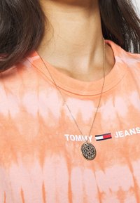 Camiseta de tirantes en naranja tie-dye con el texto blanco "TOMMY JEANS", combinada con un collar de colgante dorado que presenta un diseño circular intrincado.