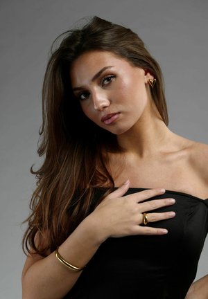 Guld smykker inkluderer hjerteformede øreringe, en ring og et glat armbånd båret af en model i en sort strapless top mod en grå baggrund.