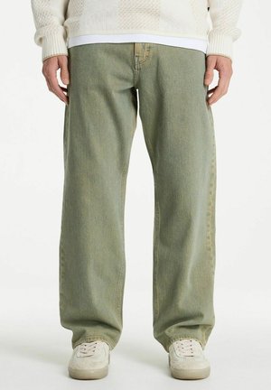 Person trägt lockere hellgrüne Jeans, weiße Turnschuhe und einen cremefarbenen Strickpullover und steht vor einem einfarbigen Hintergrund.