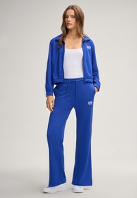 Tuta blu composta da giacca con zip e dettagli a strisce bianche, abbinata a un paio di pantaloni larghi con vita regolabile tramite cordino.