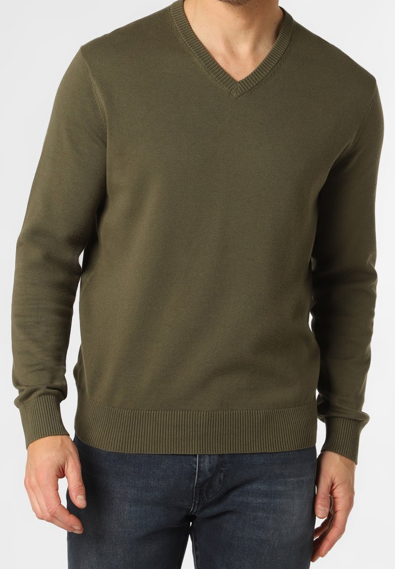 Homme portant un pull col V vert olive avec poignets et ourlet côtelés, assorti à un jean bleu foncé, debout devant un fond uni.