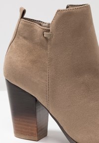 Beżowy zamszowy botek na kostkę o zaokrąglonym kształcie i obcasie typu słupek. Charakterystyczne detale to fakturowane wykończenie oraz subtelny gradient obcasa od ciemnego do jasnego.