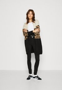 Larga chaqueta de punto en crema y estampado de leopardo, combinada con una blusa blanca, pantalones ajustados negros y zapatillas negras con suelas blancas.