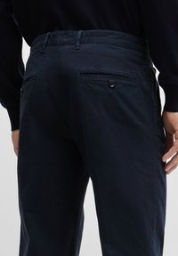 Marineblauwe broek van zacht materiaal, met achterzakken met knoopsluiting en een rechte pasvorm. Gladde textuur met een minimaal ontwerp.