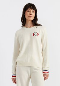 Crème-kleurige wollen trui met een Snoopy-ontwerp en rode en zwarte accenten. Geribbelde mouwen hebben rode, witte en blauwe strepen. Zachte textuur.