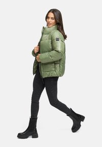 Grøn pufferjakke med høj krave, quiltet design og sort logo-patch, parret med sorte stramme jeans og klodsede sorte støvler.