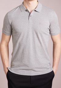 Homme portant un polo à manches courtes gris clair avec un col et des boutons, associé à un pantalon noir, les mains dans les poches.