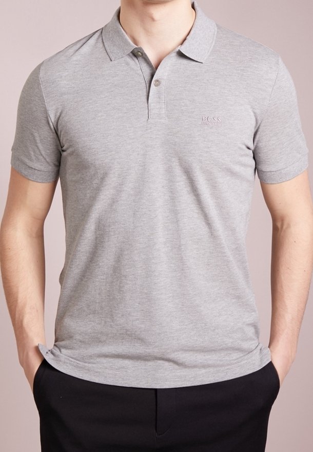 Homme portant un polo à manches courtes gris clair avec un col et des boutons, associé à un pantalon noir, les mains dans les poches.