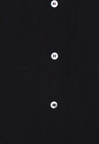 Chemise noire avec un design classique à boutons, dotée de boutons blancs et de coutures visibles. Comprend une poche poitrine. Texture lisse.