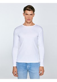 Jauns vīrietis ar īsiem brūniem matiem, valkājot vienkāršu baltu garroku kreklu un gaiši zilas džinsas, stāv pret gaiši pelēku fonu.