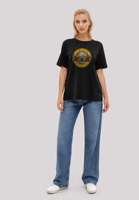Black t-shirt with graphic print of a skull surrounded by roses and text, relaxed fit, short sleeves, paired with light blue straight-leg jeans.