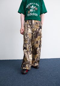 Καμουφλάζ cargo παντελόνι σε γήινες αποχρώσεις, με μεγάλες τσέπες, άνετη εφαρμογή και υφή υφάσματος. Συνδυάζεται με πράσινη μπλούζα με γραφικά.