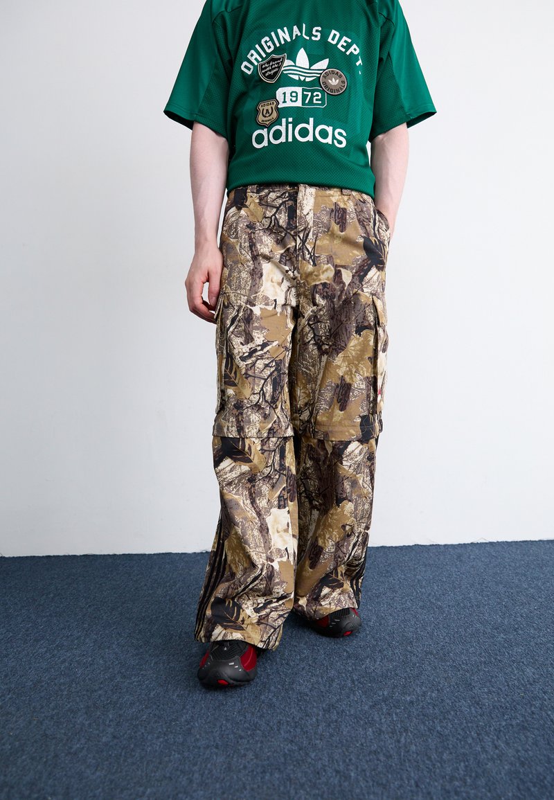 Καμουφλάζ cargo παντελόνι σε γήινες αποχρώσεις, με μεγάλες τσέπες, άνετη εφαρμογή και υφή υφάσματος. Συνδυάζεται με πράσινη μπλούζα με γραφικά.