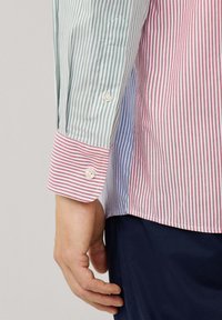 Mano al lado del torso vistiendo una camiseta a rayas multicolores con secciones en rojo, verde y azul, combinada con pantalones azul oscuro.