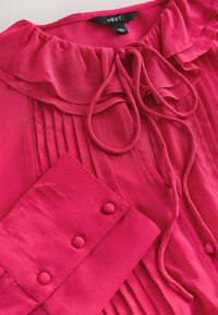 Blouse rose vif avec col à volants, liens à nouer au cou, plis verticaux sur le devant et poignets avec trois boutons recouverts de tissu.