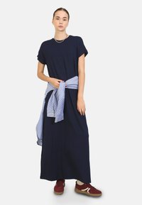 Abito maxi blu navy con maniche corte, stretto in vita con una camicia azzurro a righe legata intorno. Indossato con sneakers bordeaux.