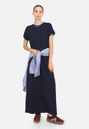 Vestido largo azul marino con mangas cortas, ajustado en la cintura con una camiseta a rayas azul clara atada alrededor. Lleva puesto unos zapatillas burdeos.