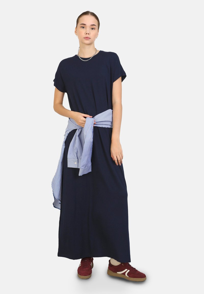 Abito maxi blu navy con maniche corte, stretto in vita con una camicia azzurro a righe legata intorno. Indossato con sneakers bordeaux.