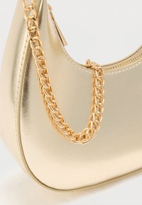 Χρυσή μεταλλική τσάντα χειρός με καμπυλωτό σχήμα, διαθέτοντας χοντρό χρυσό αλυσίδα ιμάντα και φερμουάρ κλεισίματος στο επάνω μέρος. Λείο, γυαλιστερό υφή.