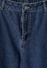 Jeans de mezclilla azul oscuro con un diseño clásico de cinco bolsillos, cierre de botón y costuras amarillas en contraste. Textura suave, sin patrones.