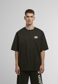 Black cotton t-shirt with short sleeves, crew neck, featuring a small, embroidered logo patch on the left side of the chest.