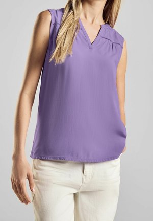 Mouwloze lavendelblouse met een V-hals, gedragen met crèmekleurige broek, rechterhand van het model in de zak tegen een effen achtergrond.