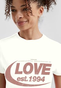 Weißes Baumwoll-T-Shirt mit einem großen roten Grafikdesign, auf dem das Wort "LIEBE", der Text "INTERNATIONALE ORGANISATION VON" und "gegr. 1994" abgebildet sind.