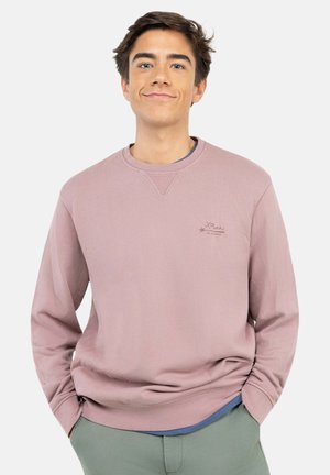 Joven con cabello castaño que lleva una sudadera morada clara y pantalones verdes, de pie con las manos en los bolsillos contra un fondo liso.
