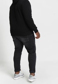 Felpa nera con cappuccio e tasca frontale, abbinata a jeans grigio scuro con taglio affusolato e leggero effetto slavato. Scarpe da ginnastica grigie con dettagli bianchi.