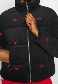 Czarna kurtka puchowa z sztruksu z wysokim kołnierzem, srebrnym zamkiem błyskawicznym oraz czerwonymi haftowanymi logo na przodzie i rękawach.