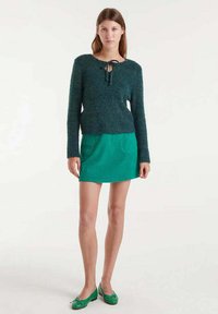 Joven mujer vestida con un suéter peludo verde oscuro con un lazo en el frente, una falda corta verde brillante con bolsillos y unos zapatos de ballet verdes, de pie en una postura neutral.