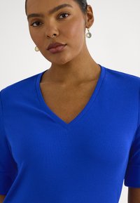 Žena so stredným odtieňom pokožky, oblečená v jasne modrej tričku s výstrihom do V a zlatými kruhovými náušnicami s gravírovaným vzorom.