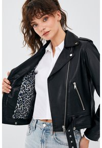 Zwarte leren motorjacket met zilveren ritsen; heeft een voering met luipaardprint. Inclusief een zijzak en drukknoopsluiting bij de taille.