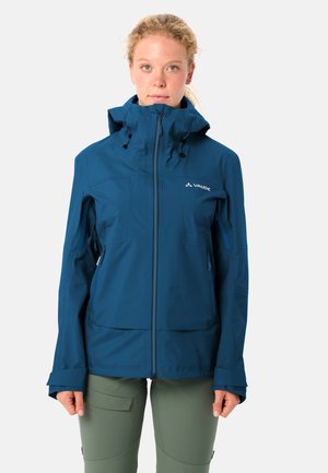 Jong persoon die een blauwe outdoorjas met capuchon en rits draagt en olijfgroene broek, staand tegen een effen lichte achtergrond.