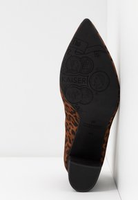 Semelle d'une chaussure à bout pointu imprimé léopard montrant des pièces en relief, la marque "KAISER" et un petit talon bloc sur fond blanc.