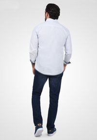 Camisa de manga larga azul claro con puños enrollados y cuello abotonado; combinada con jeans oscuros y zapatillas navy, vista desde la espalda.