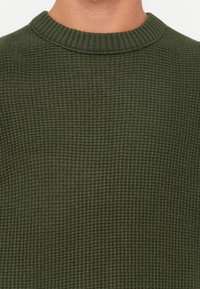 Primo piano di una persona che indossa un maglione a rete di colore verde scuro con un collo a girocollo a coste.