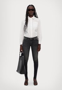 Cămașă albă cu guler, blugi skinny închiși la culoare, geantă neagră și sandale negre cu siret; model cu păr lung și ochelari de soare stă în fața unui fundal deschis.