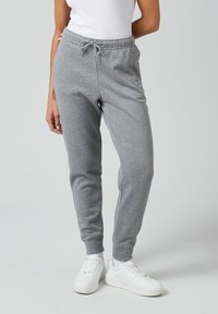 Graue Baumwoll-Jogginghosen mit einem Kordelzugbund, zulaufenden Beinen und gerippten Bündchen, kombiniert mit weißen Sneakern auf einem neutralen Hintergrund.