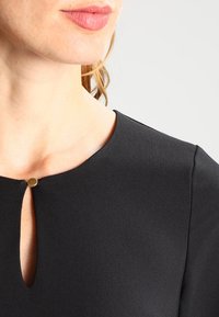 Närbild av en kvinna i en svart topp med rund halsringning och en liten nyckelhålsöppning som är fastst älld med en enda guldklick.