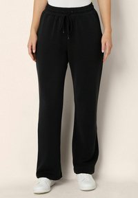 Černé bavlněné tepláky s p áskem na stažení, bočními kapsami, zúženými nohavicemi a hladkou texturou, doplněné bílými teniskami.