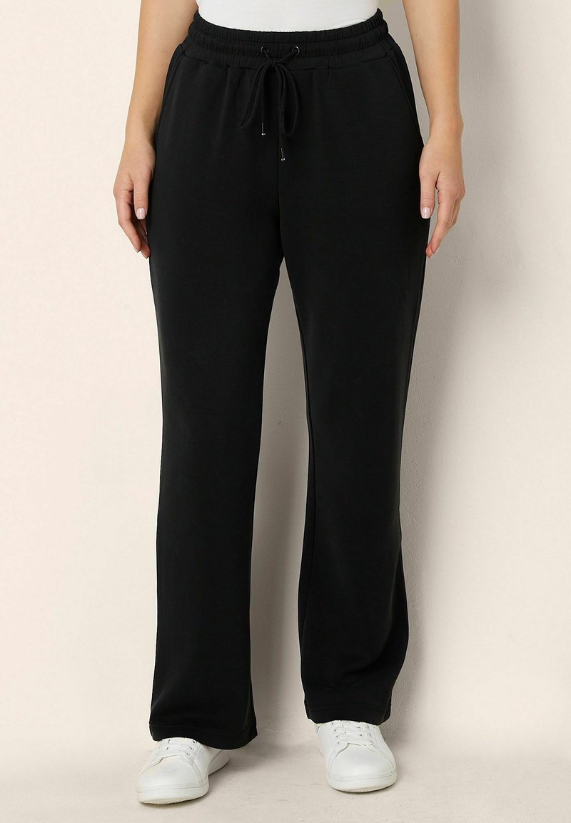 Černé bavlněné tepláky s páskem na stažení, bočními kapsami, zúženými nohavicemi a hladkou texturou, doplněné bílými teniskami.