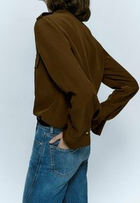 Brown long-sleeve blouse with a relaxed fit, featuring buttoned cuffs and side pockets, paired with high-waisted light blue denim jeans.