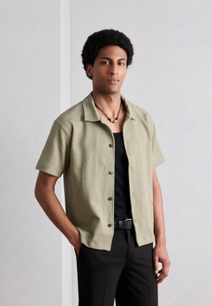 Lühikeste lokkis juustega mees, kellel on seljas beež lühikeste varrukatega nööpidega särk musta topi peal, mustad püksid ja helmestega kaelakee.