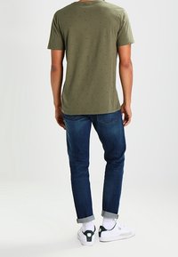 Olivengrøn t-shirt med korte ærmer og små trykte mønstre, parret med mørkeblå jeans med opkrøllede manchetter og hvide sneakers med grønne accenter.