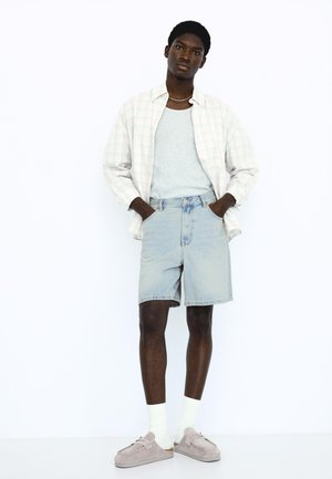 Tânăr purtând un maiou gri deschis, o cămașă în carouri deschisă, pantaloni scurți ușori din denim, șosete albe și pantofi slip-on bej, stând cu mâinile în buzunare.