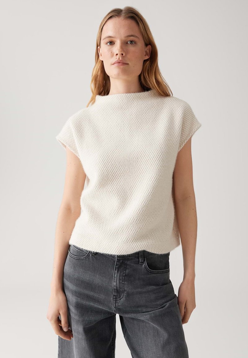 Jonge vrouw met lichtbruin haar, gekleed in een witte mouwloze top met textuur en donkergrijze jeans, staand tegen een effen achtergrond.