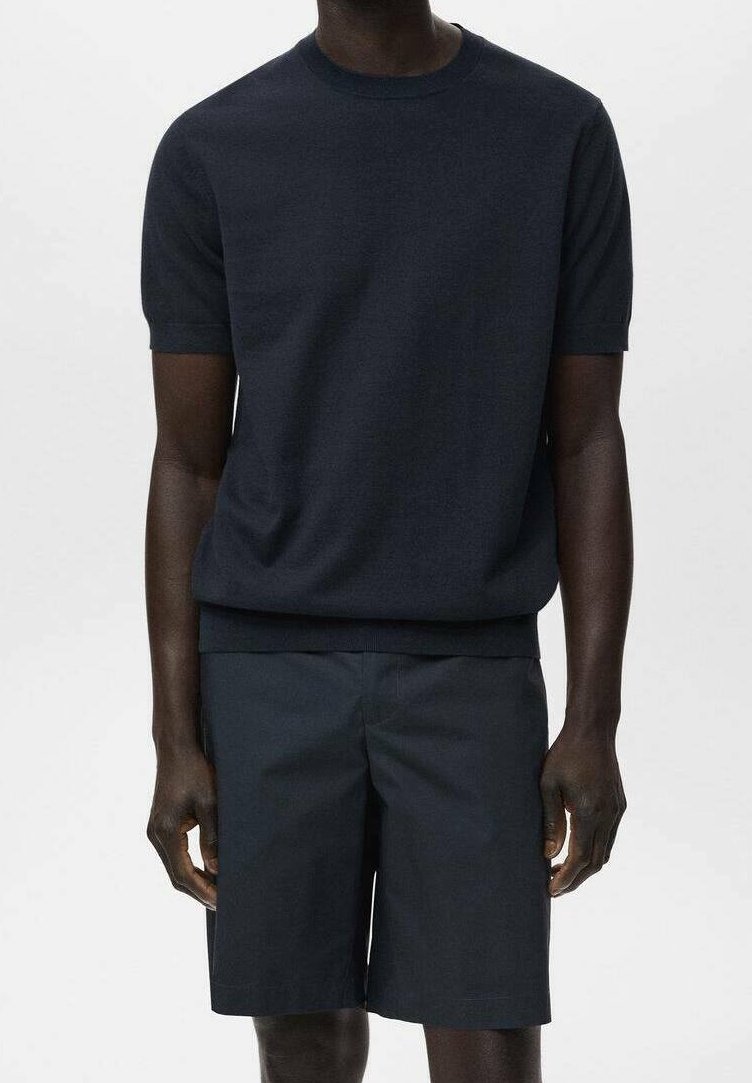Homme portant un pull à manches courtes bleu marine foncé et un short assorti jusqu’aux genoux, debout devant un fond uni.