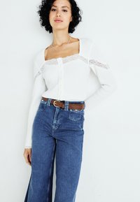 Top bianco a maniche lunghe con dettagli in pizzo e chiusura con bottoni, abbinato a jeans in denim blu a vita alta e una cintura marrone.