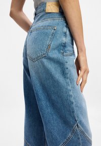 Ανοιχτό μπλε τζιν παντελόνι ψηλής μέσης με φαρδύ πόδι, διαθέτει δύο πίσω τσέπες και χαρακτηριστικές πορτοκαλί ραφές. Υφασμάτινη υφή.